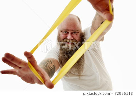 A smiling overweigh man stretches a yellow fitness elastic band and looks through it . Portrait isolated on a white background A smiling overweigh man stretches a yellow fitness elastic band and looks through it . Portrait isolated on a white background 72956118