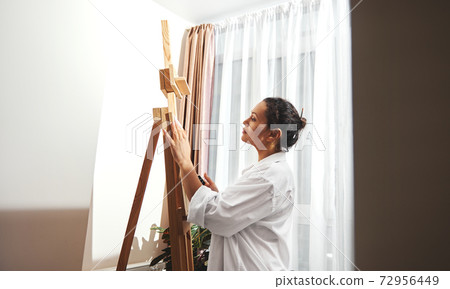 Side view of a beautiful brunette painter with gathered hair in a bun and paint brushes in her hair standing in front of the easel in workshop and drawing Side view of a beautiful brunette painter with gathered hair in a bun and paint brushes in her hair standing in front of the easel in workshop and drawing 72956449