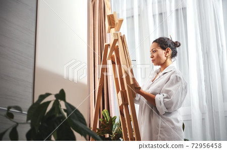 Beautiful woman painter with gathered hair in a bun and paint brushes in her hair standing in front of the easel in workshop Beautiful woman painter with gathered hair in a bun and paint brushes in her hair standing in front of the easel in workshop 72956458