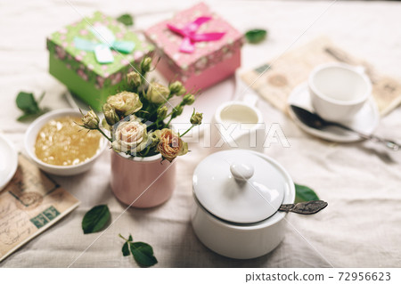 Food still life in french style. White coffee cups, milk jug with cream, roses and two gifts on a linen tablecloth. Refined toning. 72956623