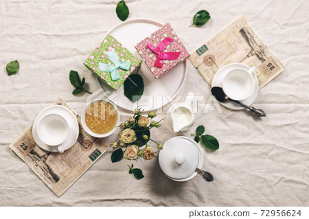 Food still life in french style. White coffee cups, milk jug with cream, roses and two gifts on a linen tablecloth. Refined toning. Top view 72956624