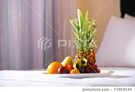 Close up of fruit plate with pineapple on white bed in hotel bedroom interior background Close up of fruit plate with pineapple on white bed in hotel bedroom interior background 72959544