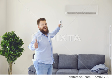Funny bearded man turning on air conditioner with remote control and showing thumb up in apartment, copy space 72961700
