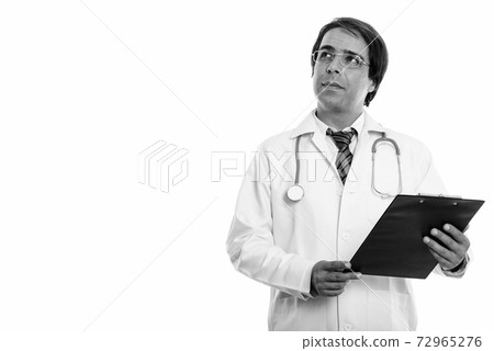 Studio shot of young Persian man doctor holding clipboard while thinking 72965276