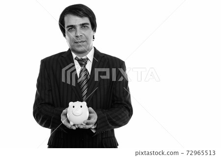 Studio shot of young Persian businessman holding piggy bank Studio shot of young Persian businessman holding piggy bank 72965513