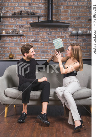 Young man gives christmas gift to his girlfriend. Happy couple of lovers at christmas eve sitting on couch in living room. New year celebration 72966281