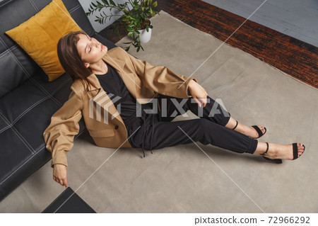 Stylishly dressed young caucasian woman in beige blazer and black pants sitting on the floor at sofa. Interior fashion portrait Stylishly dressed young caucasian woman in beige blazer and black pants sitting on the floor at sofa. Interior fashion portrait 72966292