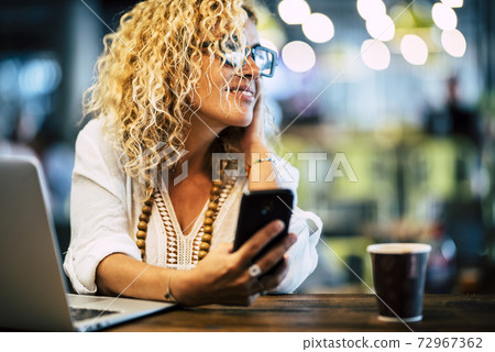 Adult blonde woman smile and work with roaning and internet technology with phone and laptop computer - office desktop workstation with defocused bokeh coloured background Adult blonde woman smile and work with roaning and internet technology with phone and laptop computer - office desktop workstation with defocused bokeh coloured background 72967362