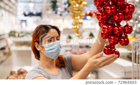 A woman in a mask chooses balls of decorations for a Christmas tree in the store A woman in a mask chooses balls of decorations for a Christmas tree in the store 72968883