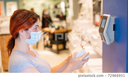 A woman in a medical mask scans the price of an item in a store. An electronic price checker on the wall in a hypermarket 72968931