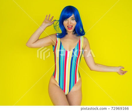 Beautiful woman in a striped swimsuit in a blue wig posing on a yellow background. Portrait of a glamorous girl. Beautiful woman in a striped swimsuit in a blue wig posing on a yellow background. Portrait of a glamorous girl. 72969166