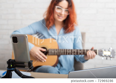 An attractive red-haired woman blogs on music. The girl sings, plays the guitar and records video on a smartphone An attractive red-haired woman blogs on music. The girl sings, plays the guitar and records video on a smartphone 72969286