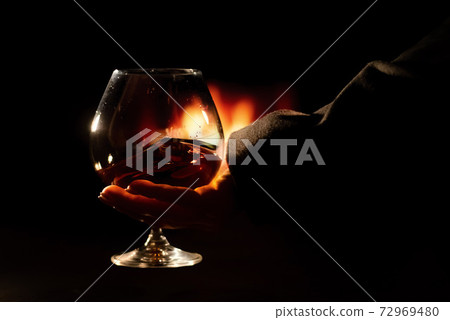 Close-up of a female hand with a glass of brandy on the background of the fireplace Close-up of a female hand with a glass of brandy on the background of the fireplace 72969480