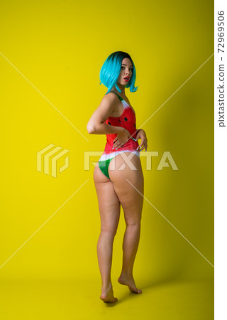 Beautiful woman in a swimsuit with a picture of a watermelon is standing with her back. Stylish girl in a colored short wig posing in the studio on a yellow background. Beautiful woman in a swimsuit with a picture of a watermelon is standing with her back. Stylish girl in a colored short wig posing in the studio on a yellow background. 72969506