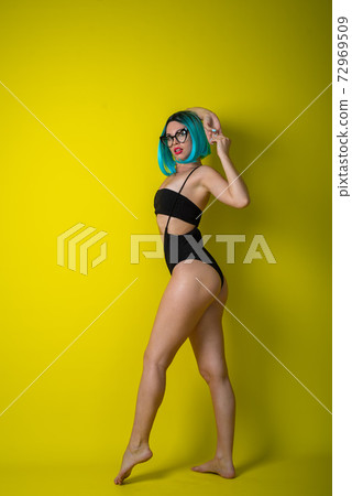 Beautiful woman in a black bikini in a colored wig and sunglasses on a yellow background. Girl posing in the studio. Beautiful woman in a black bikini in a colored wig and sunglasses on a yellow background. Girl posing in the studio. 72969509
