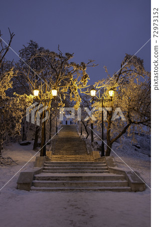 Beautiful view of the night winter city park. A ladder in the snow goes down. Lanterns by the road illuminate the path. Background for a greeting card. Beautiful view of the night winter city park. A ladder in the snow goes down. Lanterns by the road illuminate the path. Background for a greeting card. 72973152