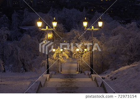 Beautiful view of the night winter city park. A ladder in the snow goes down. Lanterns by the road illuminate the path. Background for a greeting card. 72973154