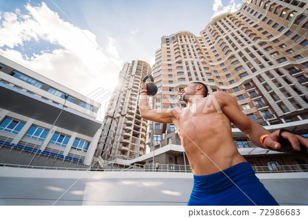 Sportive guy training with kettlebell on the background of a tall building. Sportive guy training with kettlebell on the background of a tall building. 72986683