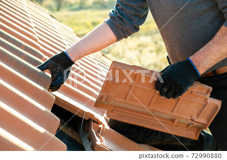 Closeup of worker hands installing yellow ceramic roofing tiles mounted on wooden boards covering residential building roof under construction. 72990880