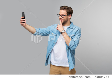 Smiling young man wearing jeans shirt taking selfie photo on smartphone or making video call standing over grey studio background 73004754