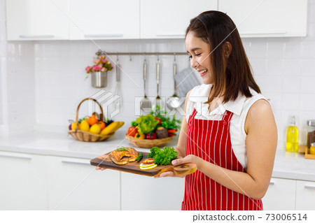 Asian woman chef in red apron holding salmon steak, serving on wooden board. Concept woman preparing meals at home. Asian woman chef in red apron holding salmon steak, serving on wooden board. Concept woman preparing meals at home. 73006514