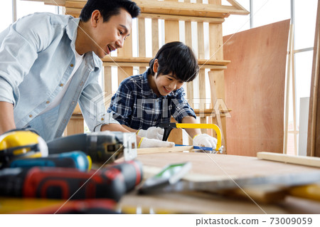 Father teach son using saw in woodwork at carpentry workshop. Concept family doing hobby together at home. Father teach son using saw in woodwork at carpentry workshop. Concept family doing hobby together at home. 73009059