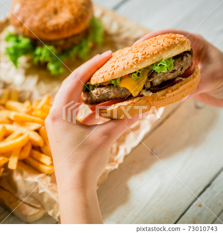 Hands and delicious burger with meat, french fries, sauce on the wooden table 73010743