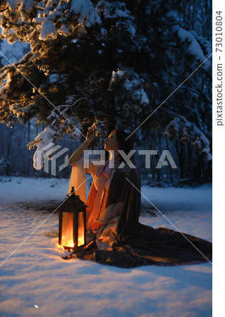 Young woman with vintage lantern sitting under fir tree covered in snow. Medieval fairytale concept 73010804