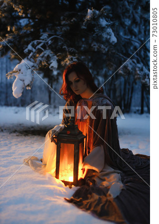 Young woman sitting under fir tree covered in snow looking at vintage lantern. Medieval fairytale concept Young woman sitting under fir tree covered in snow looking at vintage lantern. Medieval fairytale concept 73010805
