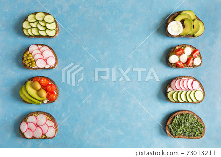 Several types homemade sandwiches with vegetables cucumbers, radishes, avocado on slices of dark bread flat lay on concrete blue background with copy space. 73013211