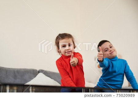 A girl in red sweater sitting on the sofa near a boy in blue sweater showing thumbs up and smiling at the camera A girl in red sweater sitting on the sofa near a boy in blue sweater showing thumbs up and smiling at the camera 73020989