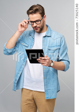 Happy young man wearing jeans shirt standing and using tablet over studio grey background Happy young man wearing jeans shirt standing and using tablet over studio grey background 73025140
