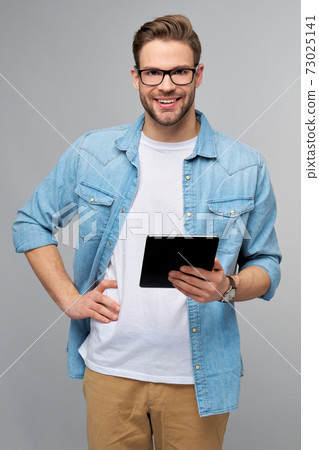 Happy young man wearing jeans shirt standing and using tablet over studio grey background Happy young man wearing jeans shirt standing and using tablet over studio grey background 73025141