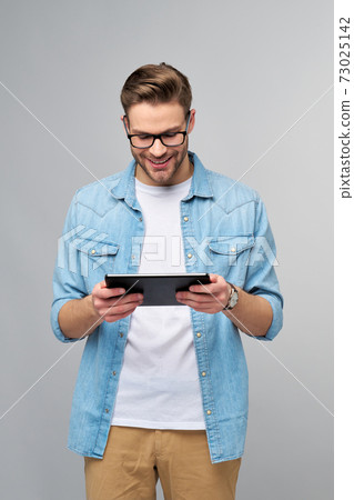 Happy young man wearing jeans shirt standing and using tablet over studio grey background Happy young man wearing jeans shirt standing and using tablet over studio grey background 73025142