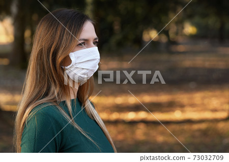 Young woman wearing white cotton virus mouth nose mask, blurred sunset lit trees in background, closeup face portrait 73032709