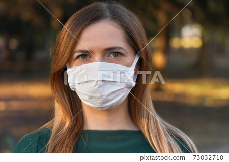Young woman wearing white cotton virus mouth nose mask, nice bokeh - blurred park trees - in background, closeup face portrait Young woman wearing white cotton virus mouth nose mask, nice bokeh - blurred park trees - in background, closeup face portrait 73032710