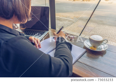 Rear view of woman writing notebook next to laptop , cappuccino Rear view of woman writing notebook next to laptop , cappuccino 73033233