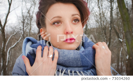 Attractive young woman with perfect skin and makeup with dark pink hair is possing on winter park background wearing blue scarf and silver earrings 73034260
