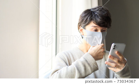 A young man wearing a mask who operates a smartphone by the window 73034367