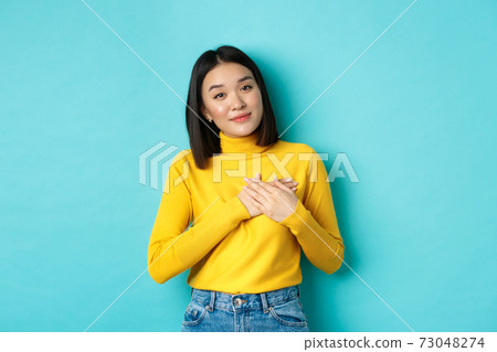 Portrait of beautiful heartfelt woman holding hands on heart, smiling and listening compassionate, standing over blue background in yellow pullover 73048274