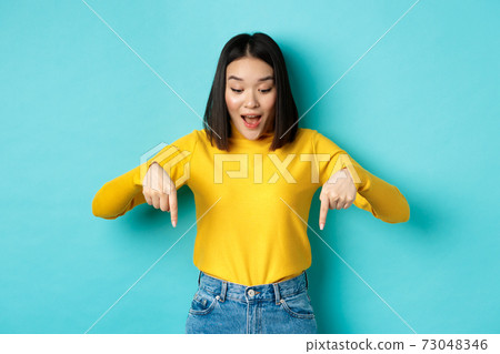 Shopping concept. Surprised cute asian girl checking out discounts, pointing fingers down and looking amazed, saying wow, standing over blue background Shopping concept. Surprised cute asian girl checking out discounts, pointing fingers down and looking amazed, saying wow, standing over blue background 73048346