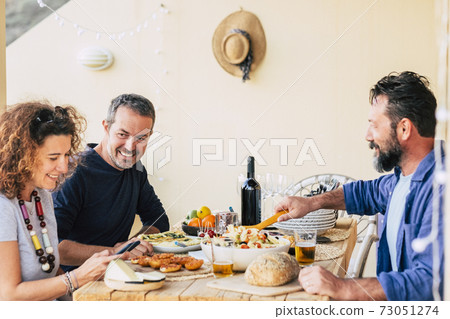 Group of adult caucasian friends enjoy lunch together at home outdoor - people enjoying food and drinks in leisure activity - friendship and cheerful people around a table with food 73051274
