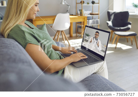 Young woman sitting on sofa with laptop and talking to online doctor of e-health clinic 73056727