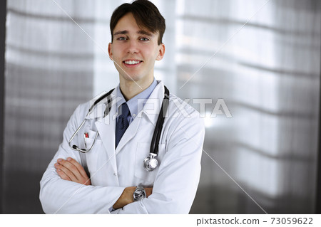 Cheerful smiling man-doctor standing with arms crossed in clinic. Perfect medical service with young smart physician in hospital. Medicine concept 73059622