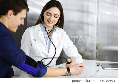 Young woman-doctor is checking her patient's blood pressure, while they are sitting together at the desk in a cabinet. Physician at work in a hospital. Medicine and healthcare concepts 73059694