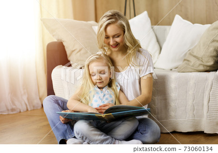 Happy family. Blonde young mother reading a book to her cute daughter while sitting at wooden floor in sunny room. Motherhood concept 73060134
