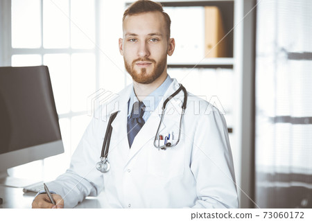 Friendly red-bearded doctor sitting and writing at clipboard in clinic. Perfect medical service in hospital. Medicine concept Friendly red-bearded doctor sitting and writing at clipboard in clinic. Perfect medical service in hospital. Medicine concept 73060172