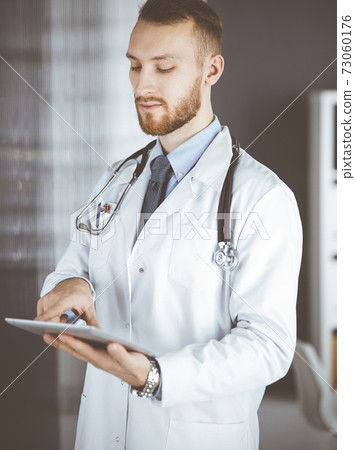 Friendly red-bearded doctor using tablet computer in clinic at his working place. Perfect medical service in hospital and medicine concept 73060176