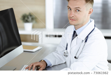 Doctor man sitting behind a computer at workplace in clinic or hospital office. Medic headshot and medicine concept Doctor man sitting behind a computer at workplace in clinic or hospital office. Medic headshot and medicine concept 73061982