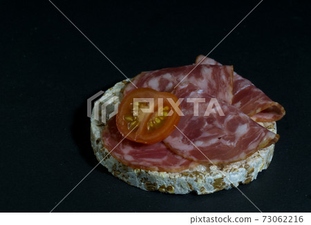 Grain crispbreads crispy rice diet bread and smoked sausage sandwich with half cherry tomato. Rice cake puffed rice with salami, sausage, balyk, smoked meat on black background Grain crispbreads crispy rice diet bread and smoked sausage sandwich with half cherry tomato. Rice cake puffed rice with salami, sausage, balyk, smoked meat on black background 73062216
